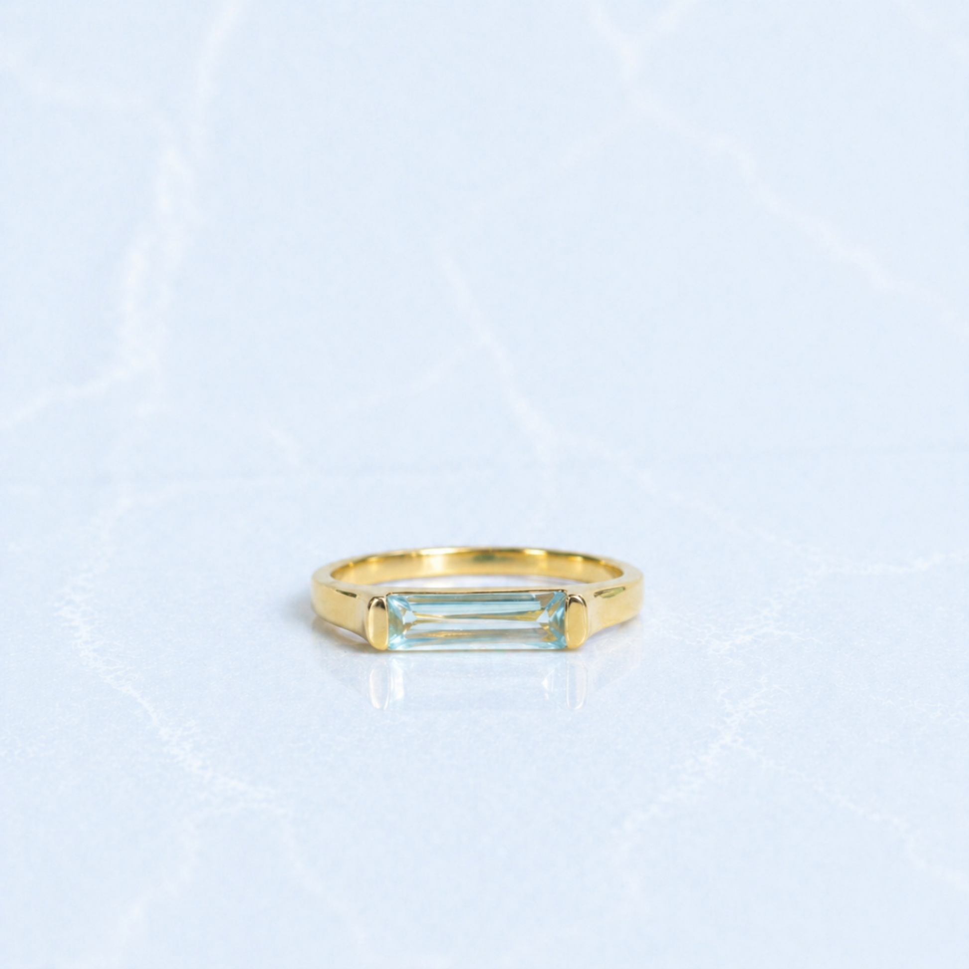 A handcrafted 18kt gold plated Sky Glow Ring featuring a rectangular blue topaz stone with a soft sky-blue hue, set in a polished minimal 925 sterling silver band on a light marbled background.
