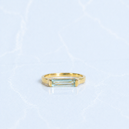 A handcrafted 18kt gold plated Sky Glow Ring featuring a rectangular blue topaz stone with a soft sky-blue hue, set in a polished minimal 925 sterling silver band on a light marbled background.