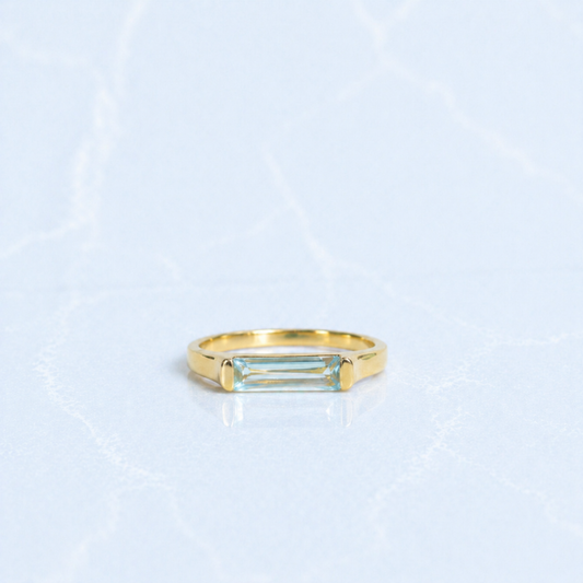 A handcrafted 18kt gold plated Sky Glow Ring featuring a rectangular blue topaz stone with a soft sky-blue hue, set in a polished minimal 925 sterling silver band on a light marbled background.