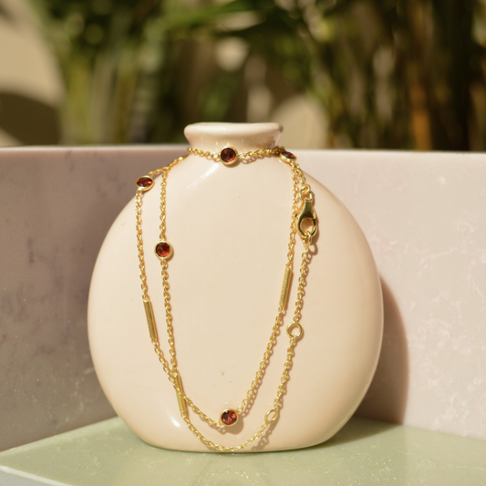 A handcrafted 18kt gold plated Warm Glow Necklace featuring circular red garnet stones and gold cylindrical bars, draped over a minimal beige ceramic vase.