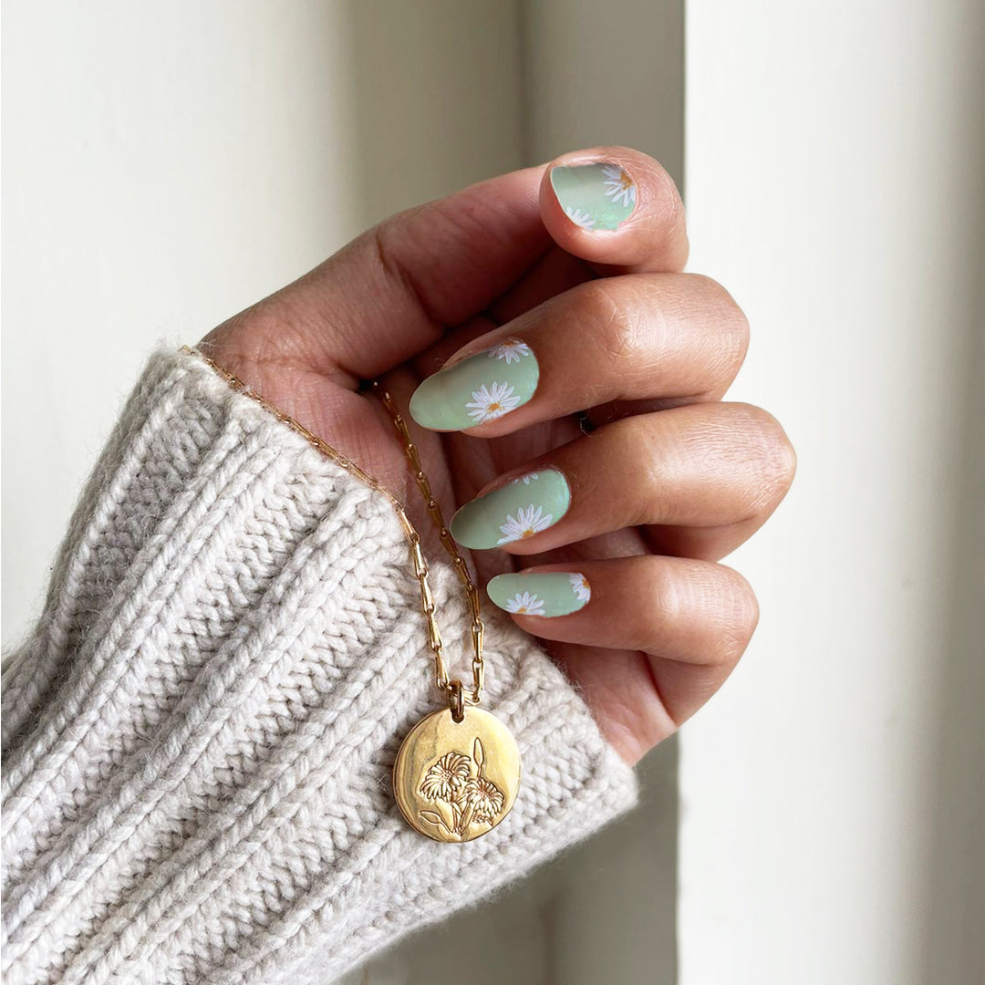 A hand holding the Daisy Medallion Necklace, featuring daisy-themed nail art that complements the floral engraving on the rose gold pendant.