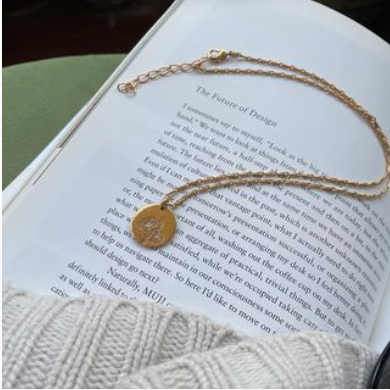 Flat lay of the Daisy Medallion Necklace resting on an open book next to a beige knit sweater, highlighting its minimalist and aesthetic appeal.