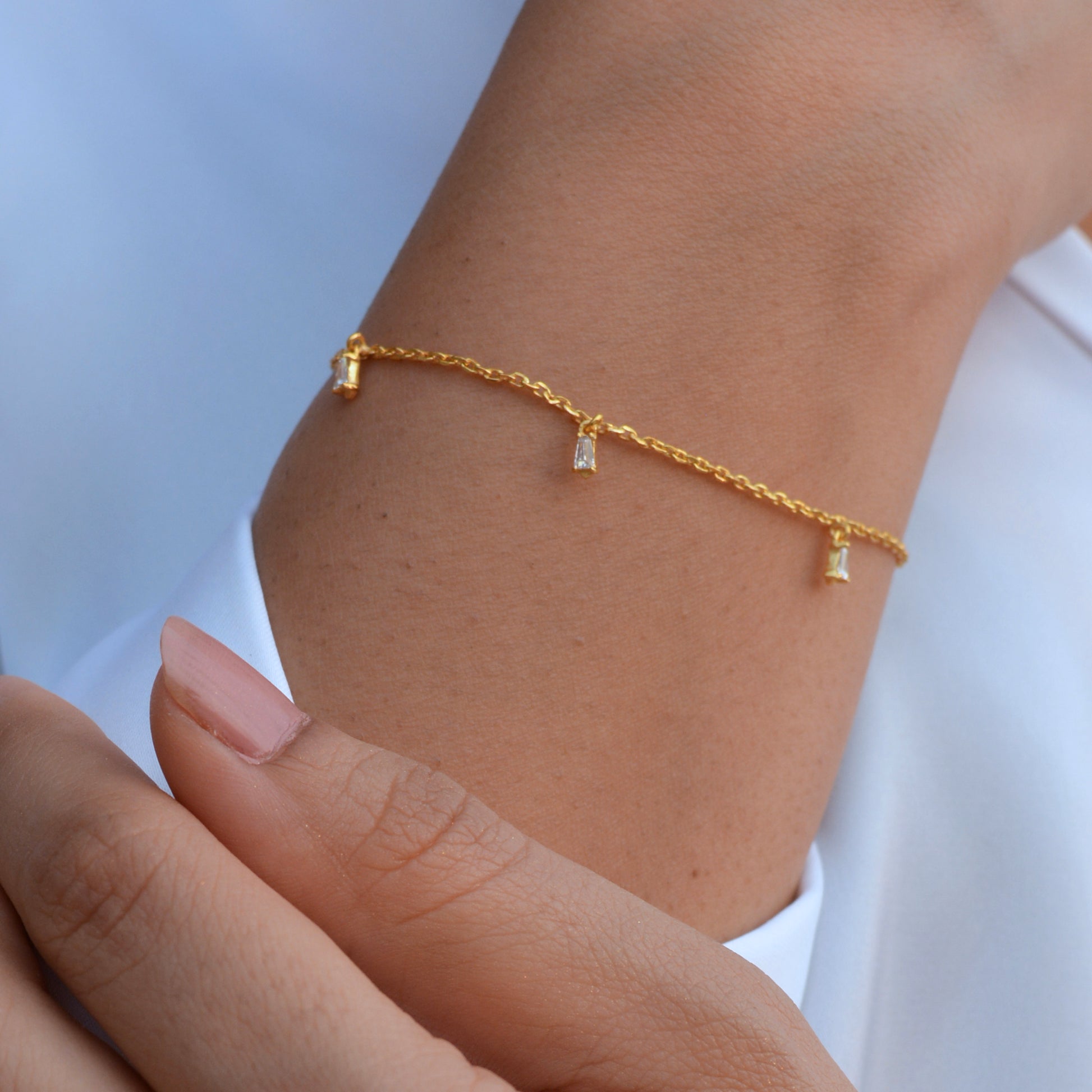 A close-up view of a person’s wrist wearing a thin gold chain bracelet with several dangling, clear baguette-shaped stones, styled against a white sleeve.