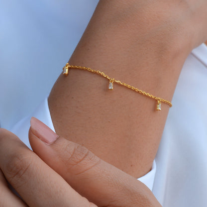 A close-up view of a person’s wrist wearing a thin gold chain bracelet with several dangling, clear baguette-shaped stones, styled against a white sleeve.