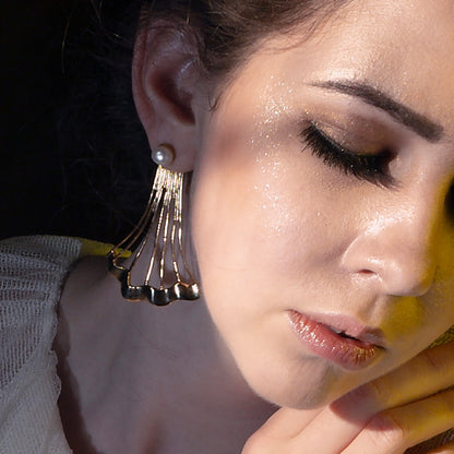Close-up of a gold fan-shaped earring with a white pearl stud worn by a model with shimmering gold makeup.
