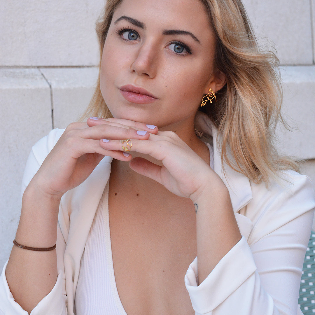 Lifestyle shot of a woman in a white blazer showcasing gold-toned botanical-inspired earrings and a thin gold statement ring.