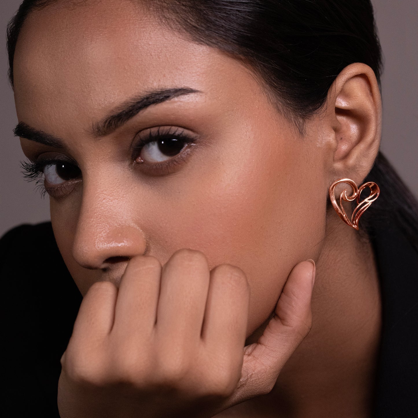 A close-up of a model's ear wearing a minimalist 18k rose gold-plated heart-shaped stud earring with a polished, high-shine finish