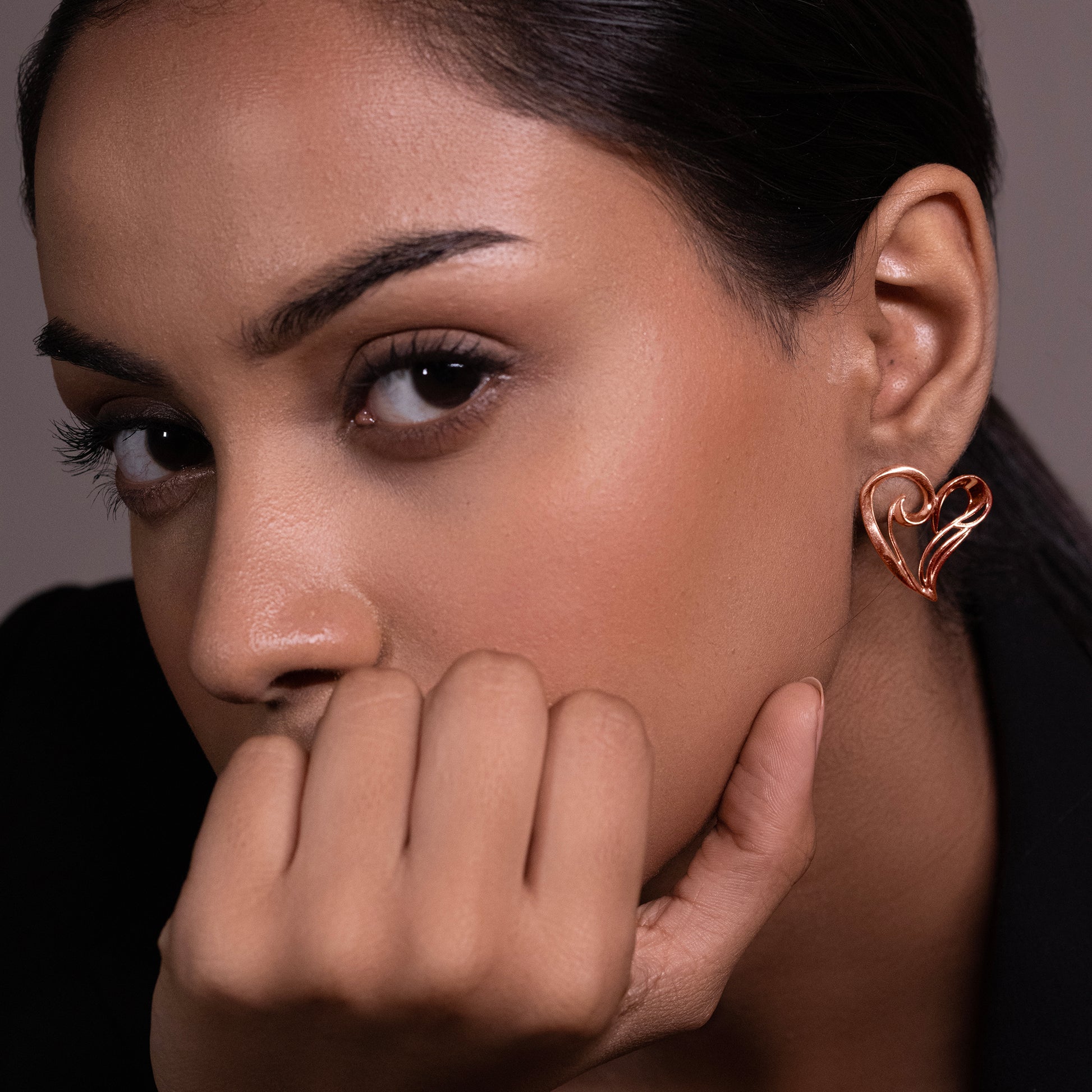 A close-up of a model's ear wearing a minimalist 18k rose gold-plated heart-shaped stud earring with a polished, high-shine finish