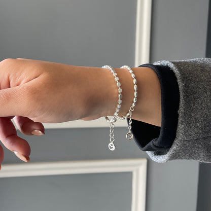 Close-up of a wrist wearing a stack of two delicate silver bracelets, one with square beads and one with oval beads, for a trendy layered look.